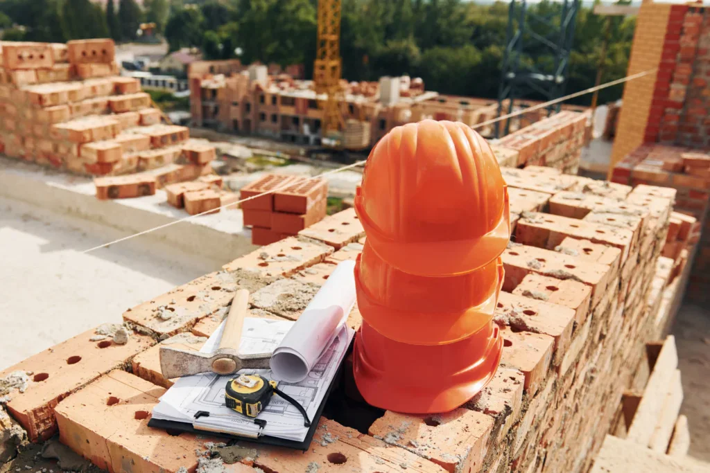 Orange hard hats, a hammer, and blueprints on a brick wall at a construction site.