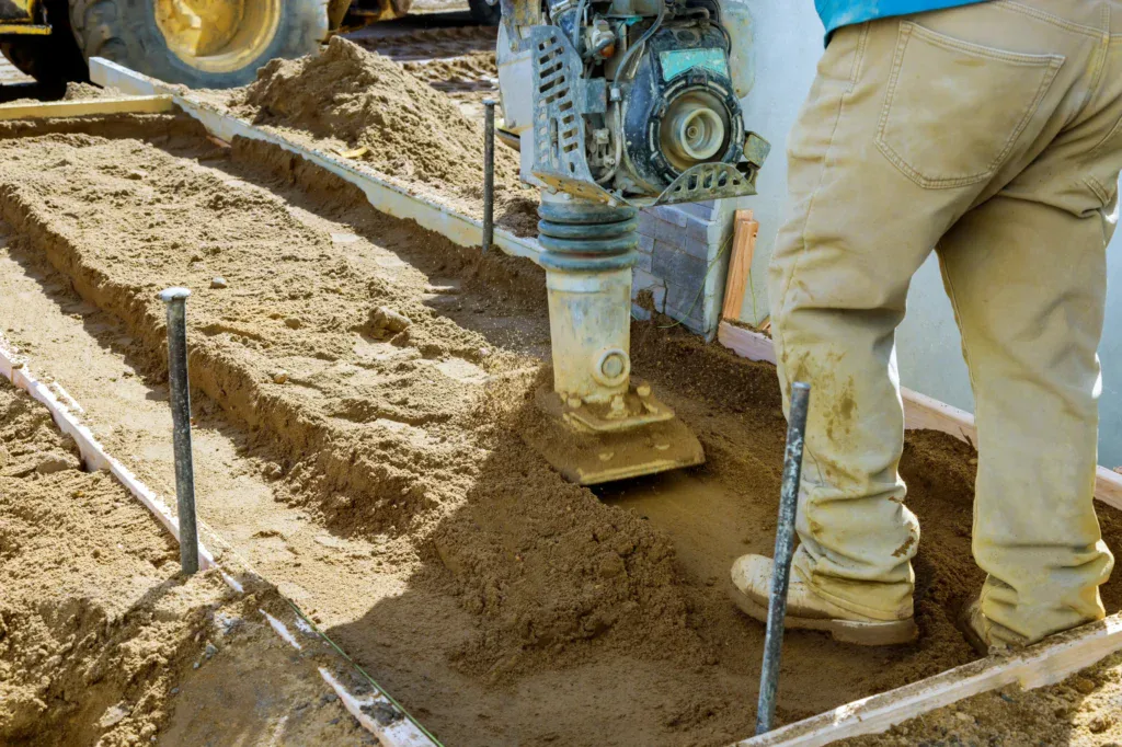 Worker compacting soil with a vibratory rammer on a construction site.