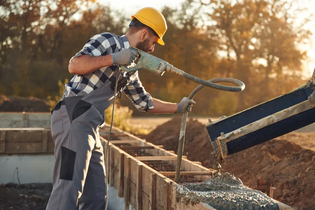 Construction worker in hard hat pouring concrete into wooden forms.