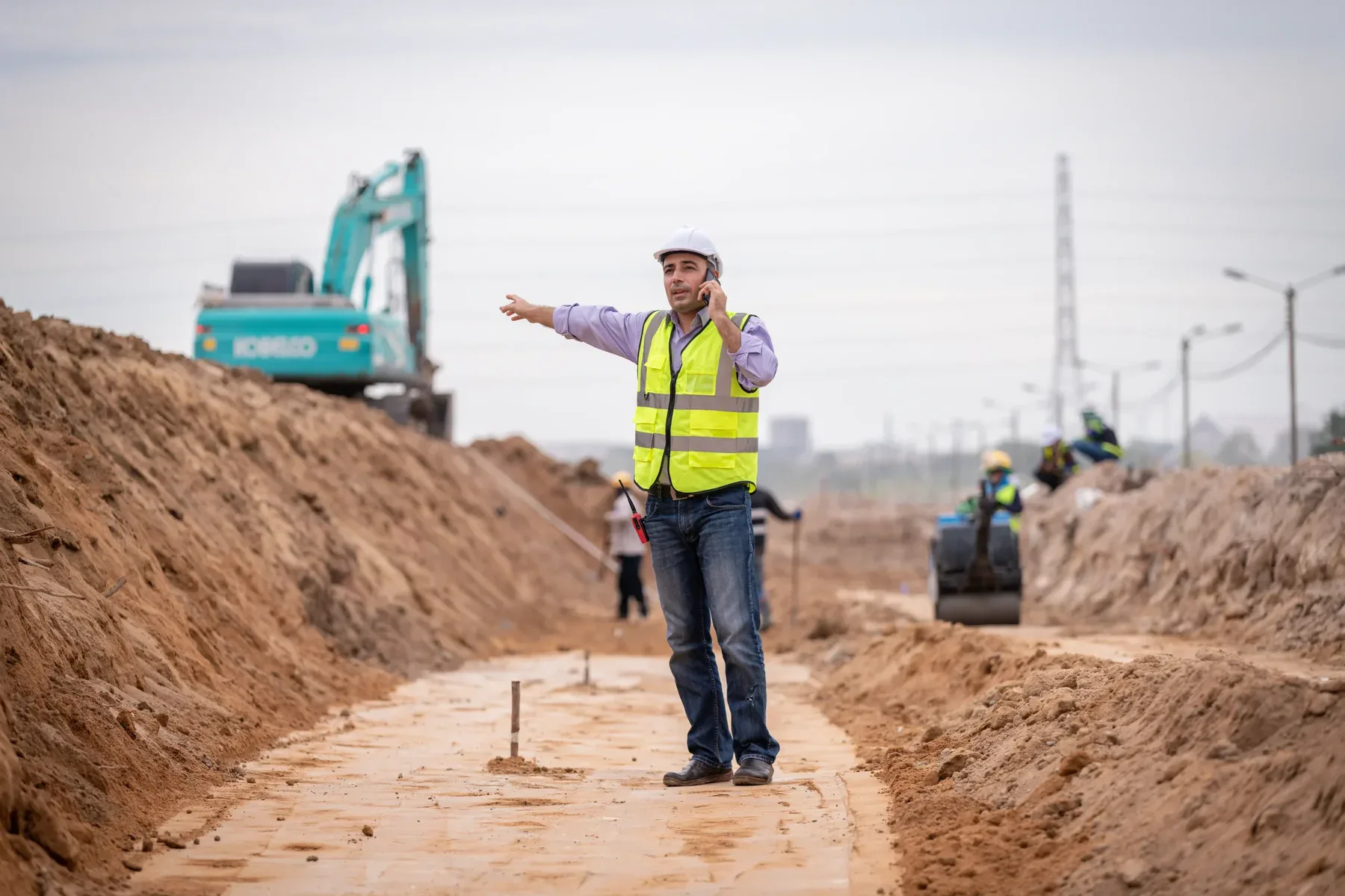 A construction worker stands on a dirt path between two excavated trenches. The worker wears a high-visibility yellow safety vest, blue jeans, and a white hard hat while gesturing with one arm extended. In the background, a turquoise excavator is positioned on the left side of the frame. The trenches are deep and contain exposed earth, creating a U-shaped corridor. The path is compacted dirt and runs through the center of the construction site. In the far background, utility poles and power lines are visible against an overcast sky. The image has a professional, documentary-style composition with natural lighting and sharp focus on the foreground subject.