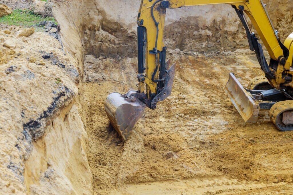 Yellow excavator digging a large trench in sandy soil.
