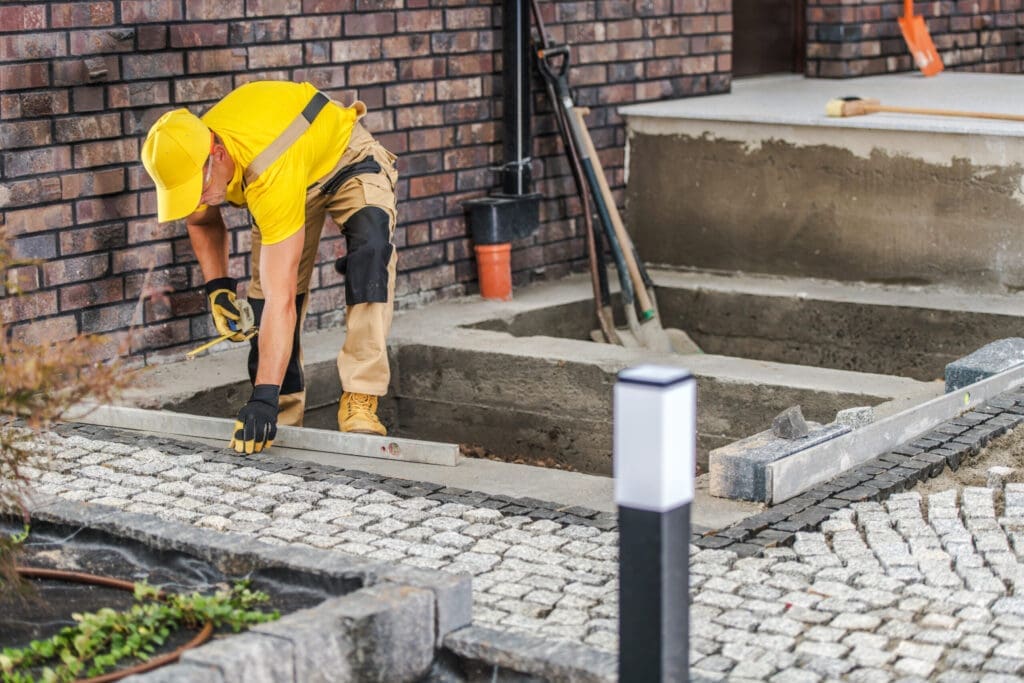 Man in yellow shirt and cap installing paving stones next to a brick wall.