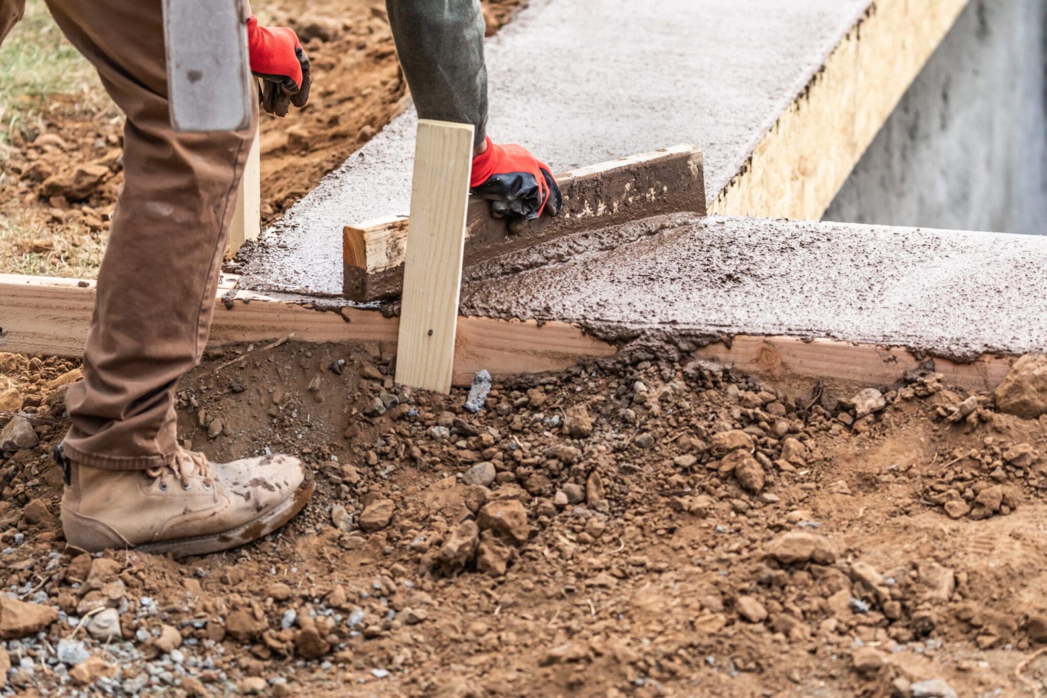 Worker in gloves and boots leveling wet concrete with a wooden tool.