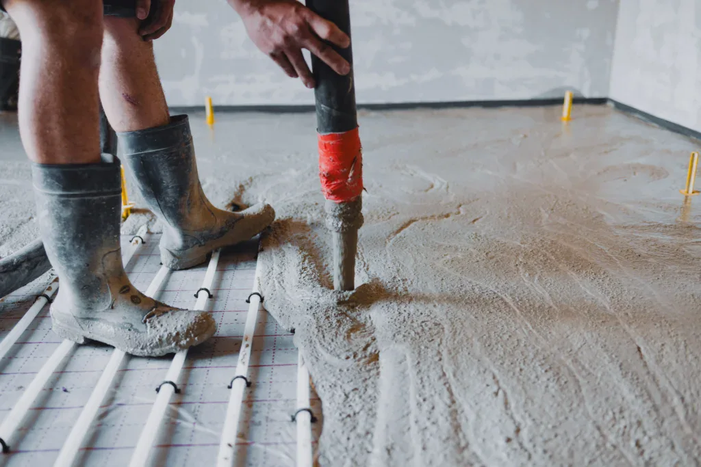 Person in boots using a tool to spread wet concrete over pipes