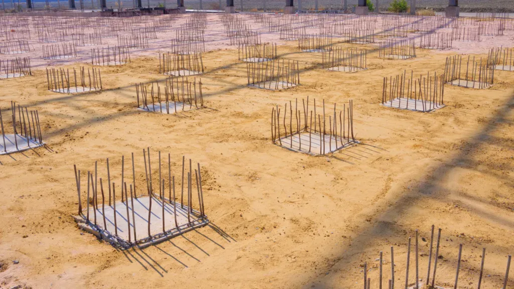Construction site with numerous concrete foundations and rebar frames on sandy ground.