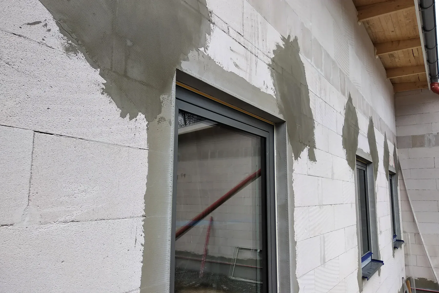 A close-up photograph of a white concrete block wall with gray stucco being applied. The wall features a modern gray-framed window installation. The stucco is being applied in sections, with three distinct patches of light gray stucco visible on the right side of the frame. The stucco application is in progress, with fresh gray material contrasting against the white concrete blocks. The window frame is a contemporary style with a dark gray finish. The ceiling visible in the frame shows exposed wooden beams. The lighting in the image is natural and even, casting subtle shadows where the stucco meets the concrete blocks. The composition is shot at a slight angle, showing the depth of the window installation and the texture of both the concrete blocks and the stucco application.