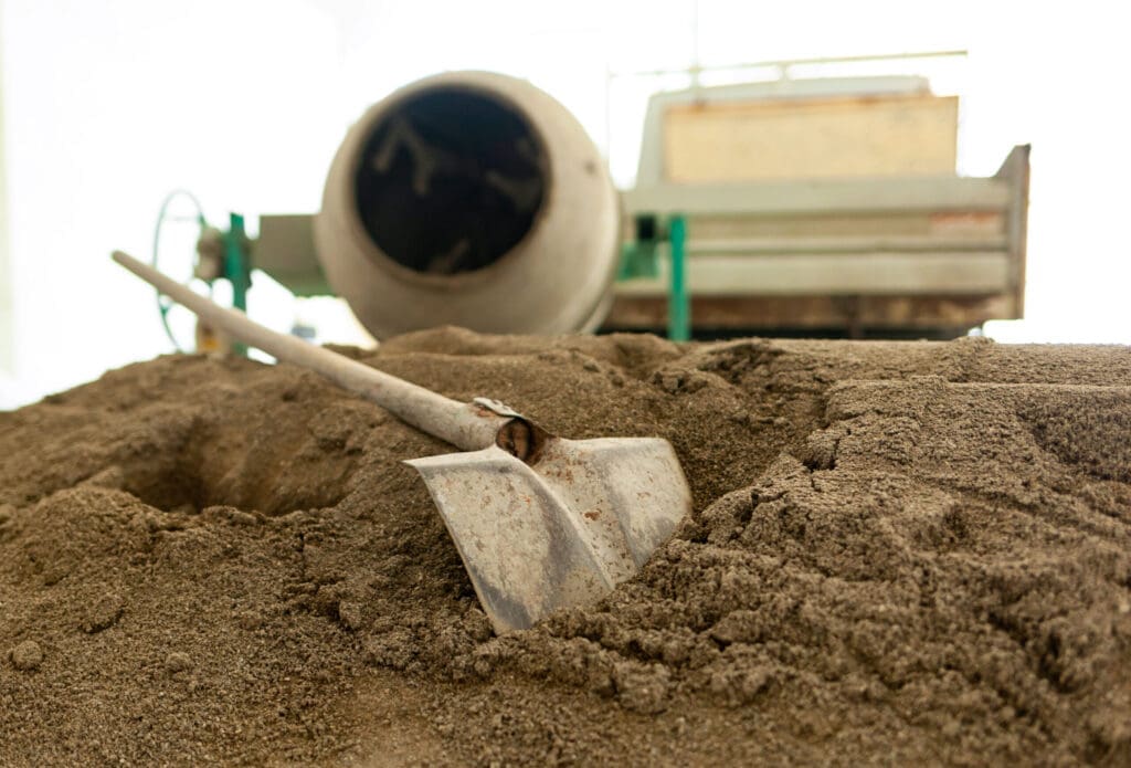 Shovel in a pile of sand with a cement mixer in the background.