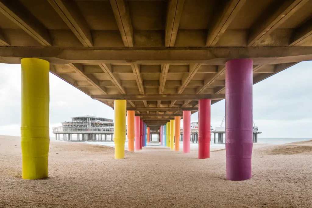 Colorful pillars under a pier on a sandy beach leading to a building.