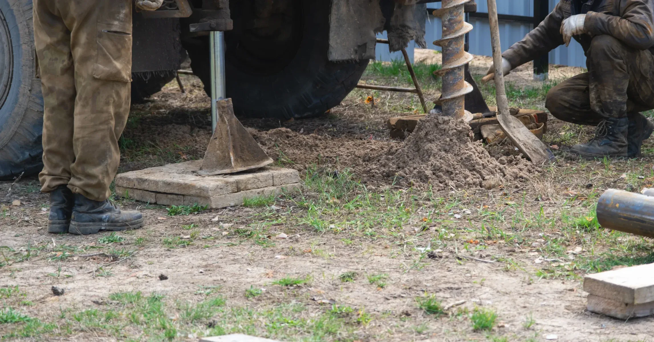 Two workers and drilling equipment on a dirt and grass patch