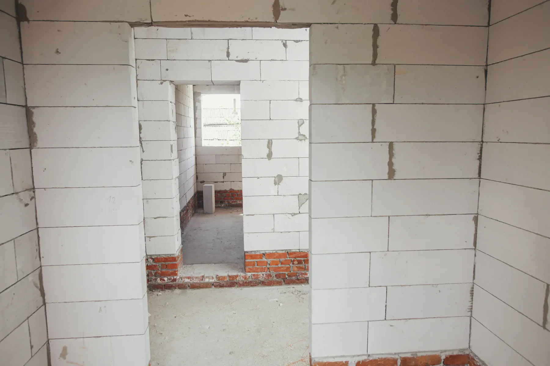 A photograph of an unfinished interior room with white concrete block walls and exposed brick foundation. The walls are constructed with large rectangular white blocks, with visible mortar lines and some patches of exposed concrete. A doorway leads to another room, where a small window lets in natural light. The floor is unfinished with exposed red brick at the base of the walls. The ceiling shows exposed structural elements and unfinished surfaces. The walls have a rough, industrial appearance with some areas showing wear and discoloration. The lighting in the image creates subtle shadows between the block walls, emphasizing the architectural details and construction materials.