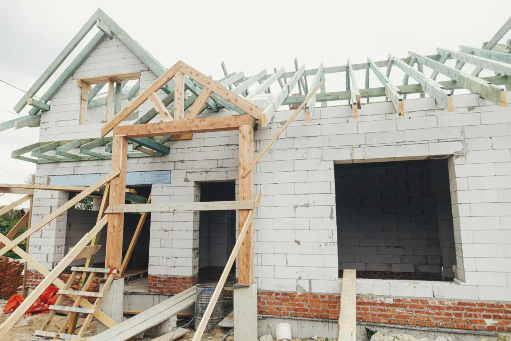 Unfinished house with white brick walls and wooden roof trusses under a cloudy sky.