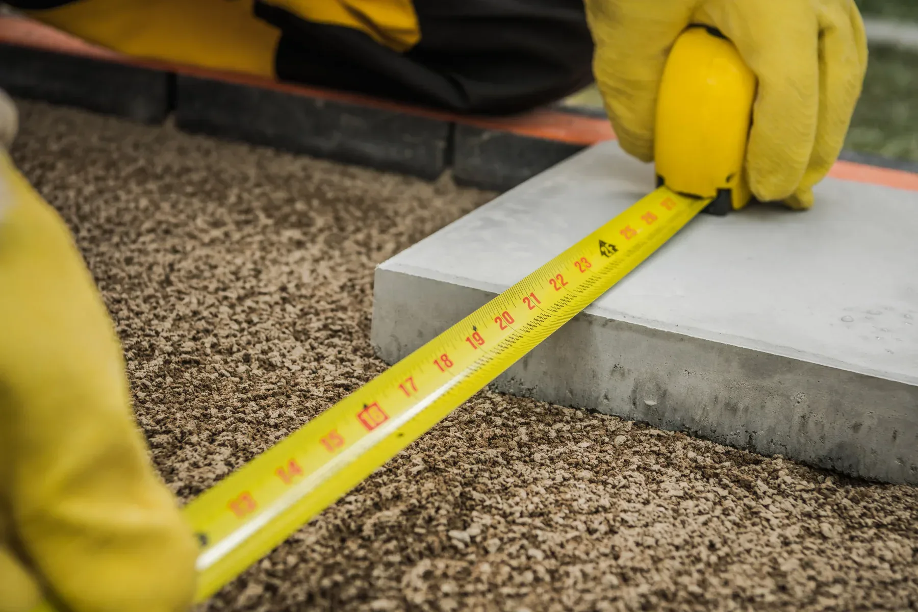A close-up photograph of hands wearing bright yellow work gloves measuring a concrete surface with a yellow measuring tape. The measuring tape has red numerical markings and extends across the frame. The concrete surface is a light gray color with a smooth finish. The background shows a textured gravel surface in a light brown color. The image is shot from above at a slight angle, with the measuring tape being held horizontally across the concrete surface. The yellow gloves are rubber or neoprene material, and only portions of the hands are visible in the frame. The lighting is even and natural, highlighting the contrast between the yellow measuring tape and the gray concrete surface.