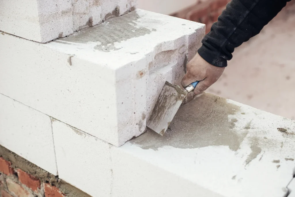 A person's hand applying mortar to white aerated concrete blocks.