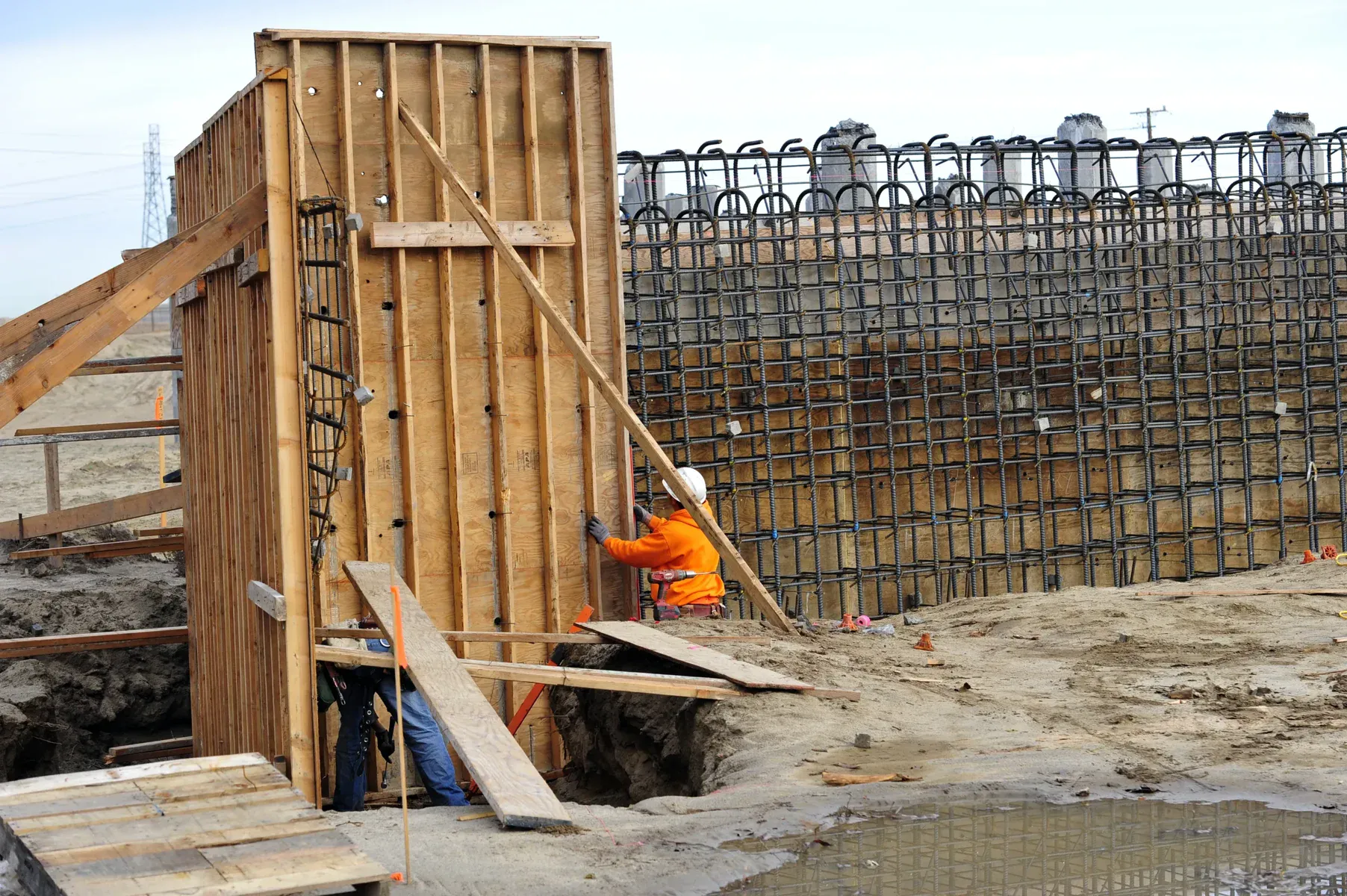 Construction worker in orange safety vest next to wooden formwork and rebar wall.
