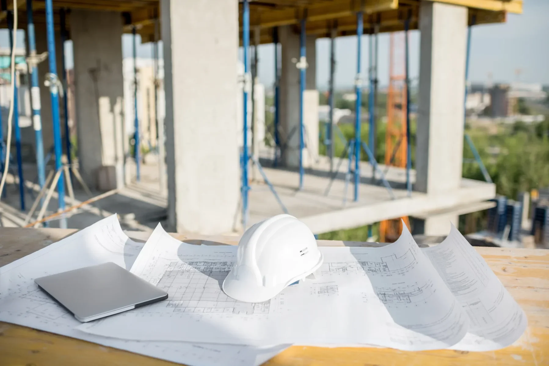White hard hat, laptop, and blueprints on a wooden surface at a construction site.