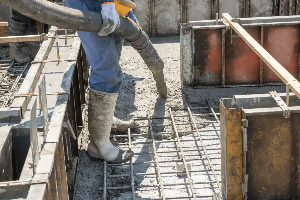 A close-up photograph of construction work in progress. A worker wearing blue jeans, a light blue long-sleeved shirt, and gray rubber boots stands on metal rebar reinforcement bars. The worker is operating a gray concrete pump hose that extends out of frame. The scene shows a concrete foundation with metal formwork panels and wooden support structures. The formwork has a reddish-brown stain on its surface. The rebar creates a grid pattern across the concrete surface, with individual steel rods visible. The lighting is natural daylight, creating subtle shadows on the concrete surface. The image is cropped to focus on the worker's midsection and the construction materials, showing the industrial nature of the concrete pouring operation.