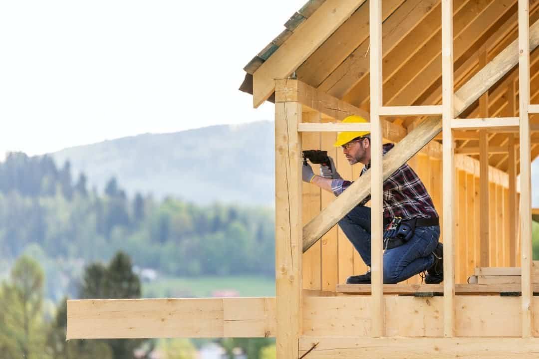 Construction worker inspecting wood framing on a new house under construction, wearing safety gear and using a level during framing inspection, with a scenic outdoor background.