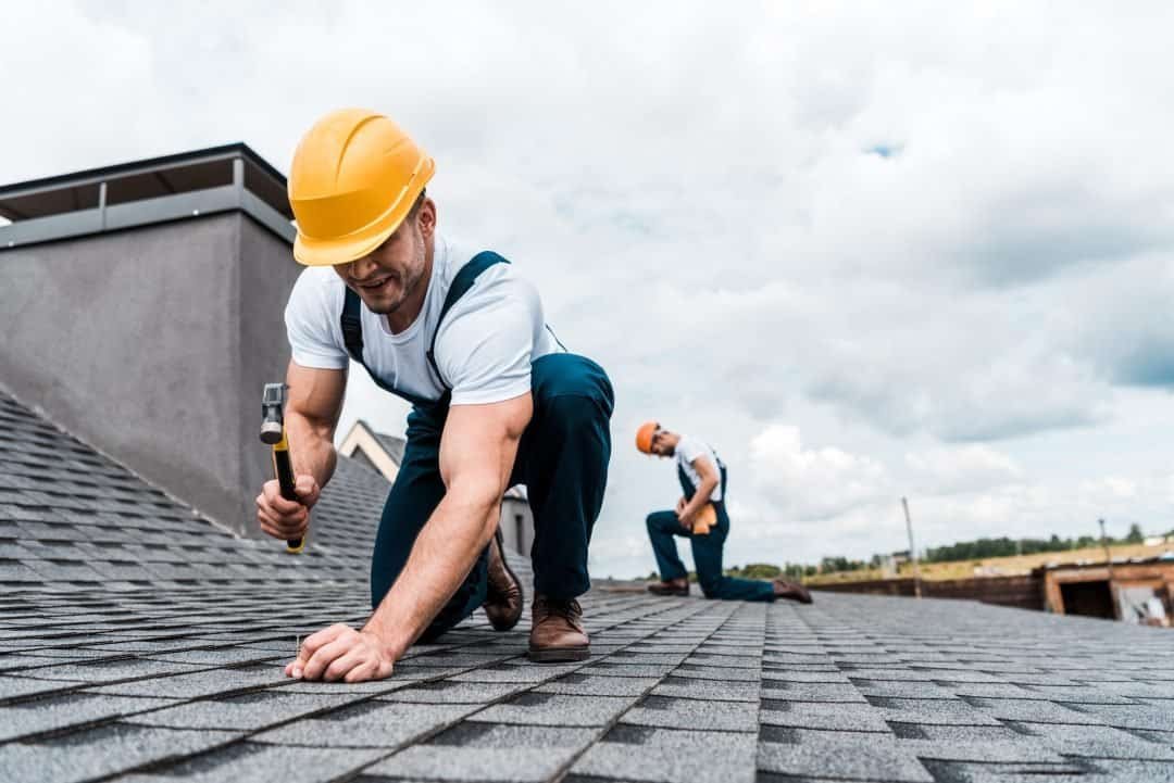 Shingle roof installation by professional construction workers wearing safety helmets on a residential building.