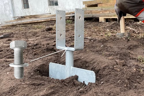 close-up outdoor shot of a metal GroundPlug® bolt bracket installed in the ground. The bracket is T-shaped with a U-shaped section at the top, and a large metal bolt lies horizontally nearby. The ground is dark, damp soil, and the background shows a building with a white wall and a stack of wooden planks. A person's gloved hand and a hammer are partially visible on the right.