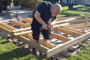 Carpenter operating an impact wrench to drive a screw pile. He is standing in between thick wooden beams, so the screw piles can be attached to the structure. He is working on an outdoor home improvement project.