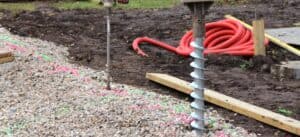 Two ground screws installed on a gravel and dirt construction site, one with a drive head attached, near coiled orange tubing.