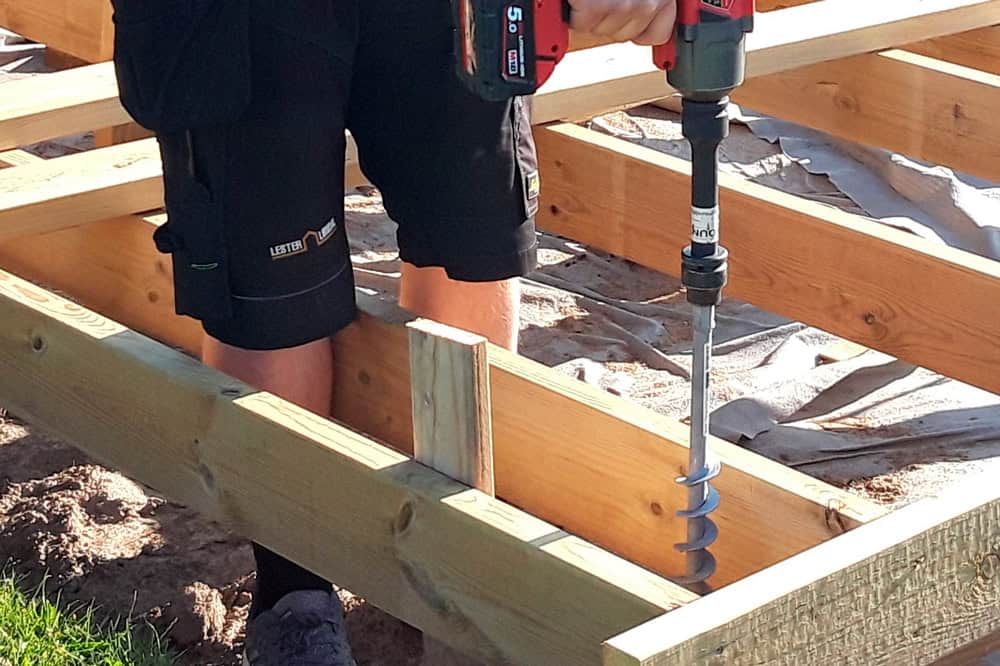 Close-up of a real-life steel GroundPlug® screw pile being drilled into soil, with a man holding an impact wrench attached to the foundation.
