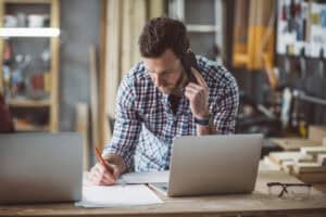 Individual manages tasks at a desk in a workshop setting, simultaneously engaging in a phone call and writing notes, using two laptops.