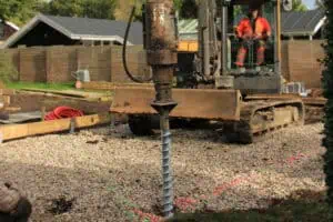 Excavator operated by a person in an orange jacket installs a ground screw on a gravel construction site.