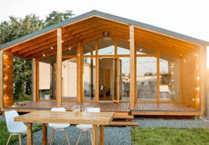 A contemporary single-story building made of natural wood, featuring a gable roof extending over a spacious deck, and a floor-to-ceiling glass front wall. String lights decorate the covered area, and a dining set sits on the gravel patio.