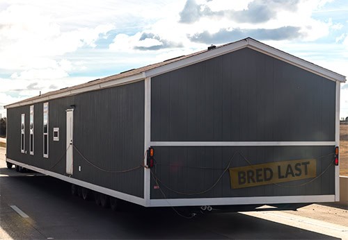 Section of a grey mobile or modular home being transported on a trailer on a highway, with a "BRED LAST" sign on the back.