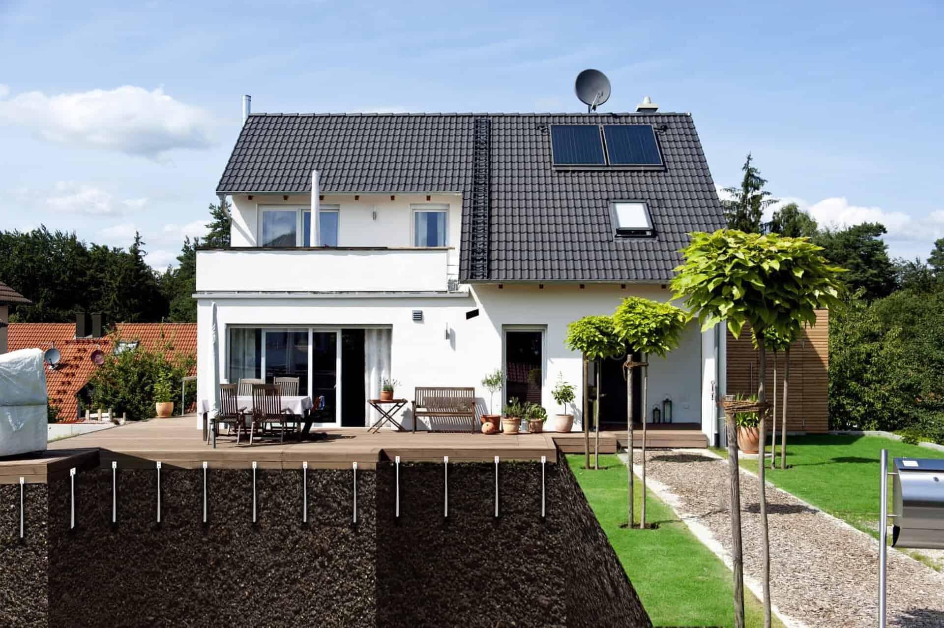 Modern white house with a dark roof, solar panels, a wooden deck, and a cutaway view showing the foundation supports in the ground.