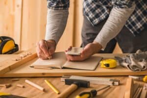Woodworker consulting a smartphone and taking notes with a pencil in a notebook laid on a workbench amidst woodworking tools and materials.