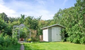 A glass greenhouse stands next to a garden shed with white doors on a green lawn, surrounded by dense trees and bushes under a blue sky with light clouds. It shows that screw pile foundations have many benefits when you want to build a shed structure.