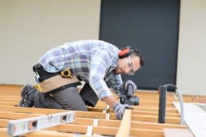 Person engaged in constructing a wooden deck frame, kneeling and using a power tool to fasten beams, while wearing protective gear. This deck is standing on screw piles, which are great for both professional and diy deck projects. What are the best decking materials out there to use with screw piles?