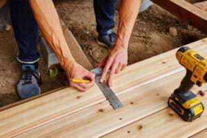 Worker measures and marks a piece of lumber with a square and pencil as part of a deck building project, with a power drill sitting on the wood.