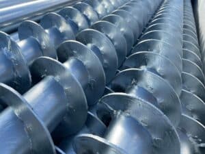 A close-up shot of PilexPro groundplug foundations screw piles arranged in a row. The piles are made of silver metal and have a helical flange design.