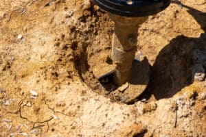 A close-up shot of a metal auger post hole digger drilling into dry, cracked earth. The auger is centered in the frame, with a hole visible around it. The earth is light brown with visible cracks and small pieces of debris. The top of the digger is partially visible at the top of the frame. The scene suggests the process of digging a hole for a post or other construction purpose. It can also suggest a screw pile being installed, providing cost-effective foundations for that type of soil.
