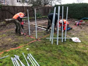 Screw piles in rain are the most efficient solution in rainy season. In the image, a man in a bright orange jacket is installing a screw pile with a red, hand-held impact wrench, while in the surrounding area there are more screw piles ready for installation.