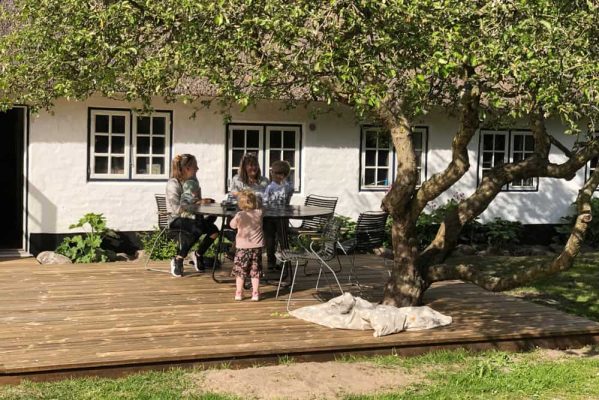 Several people, including adults and children, are gathered around a round metal table on a sunny wooden deck, shaded by the branches of a large leafy tree. The deck adjoins a white house with multi-pane windows.