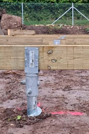 A galvanized metal ground screw installed in muddy soil, with its top bracket securely bolted to thick, stacked wooden support beams, forming part of a foundation.