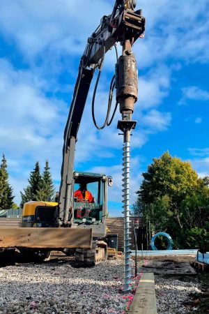 Operator using an excavator equipped with a drilling attachment to drive a helical pier foundation into a gravel worksite marked with guidelines, next to a wooden beam.