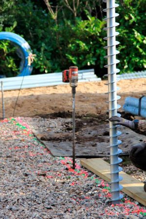 Construction site featuring a large ground screw being manually guided into place next to a wooden beam on gravel, while a separate power auger stands vertically further back.