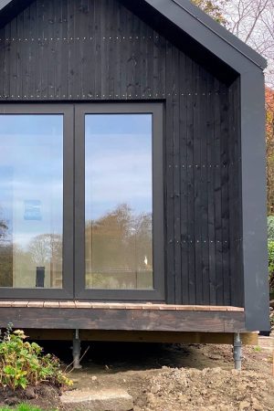 Close-up of a modern black tiny house corner with large windows, showing the elevated deck and adjustable pier foundation.