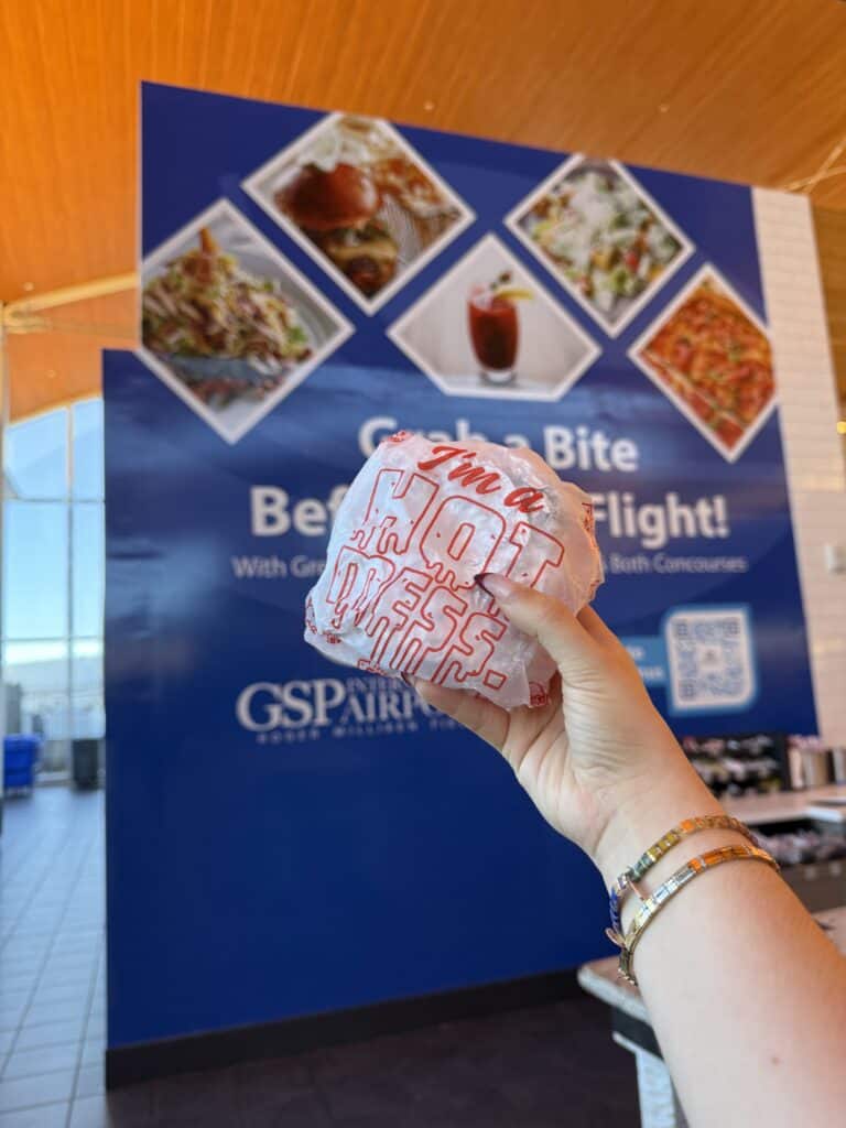 Hand holding up a sandwich wrapped in a paper in front of airport food hall.