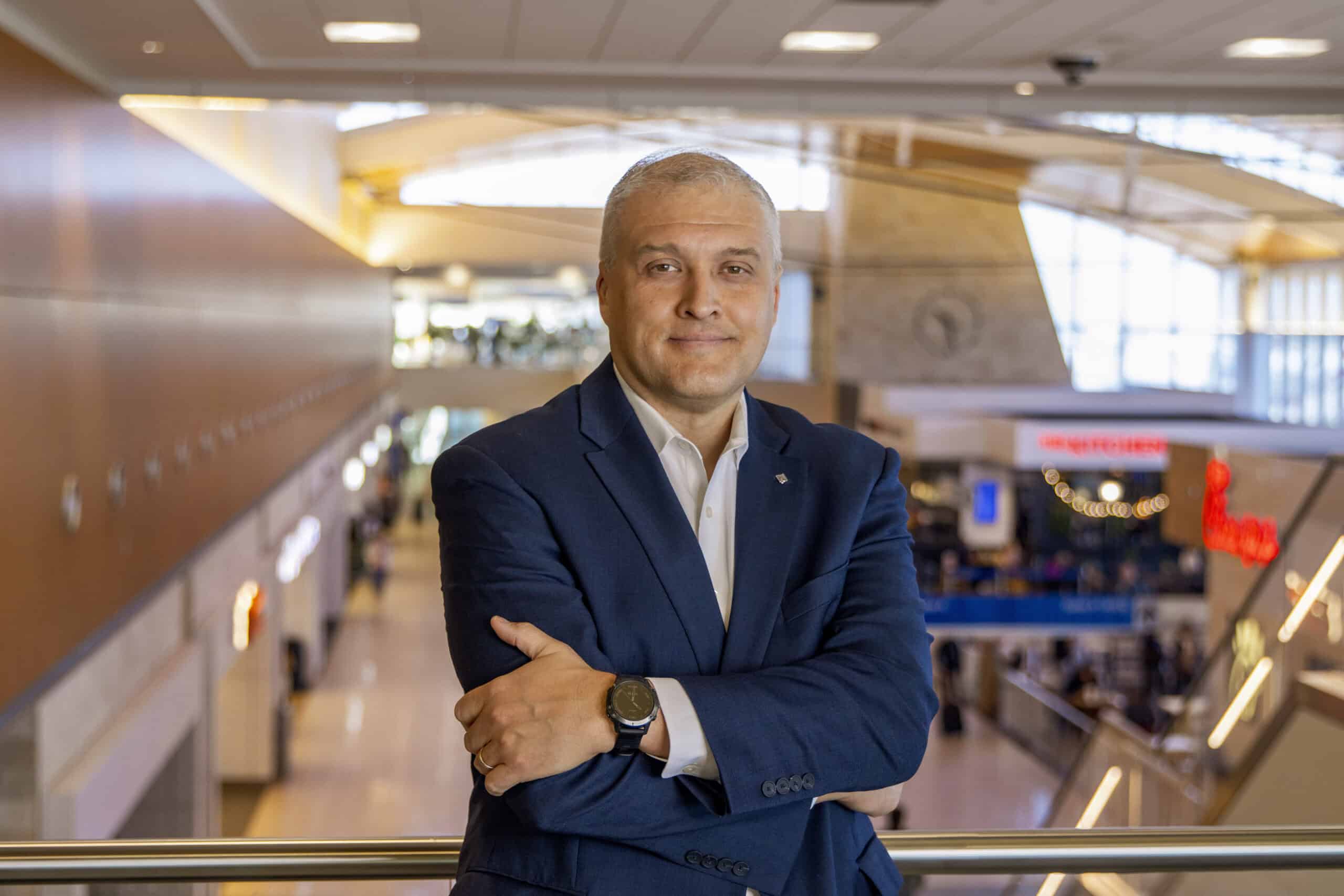 Photo of executive is a blue jacket with his arms crossed and interior of airport terminal in the background.