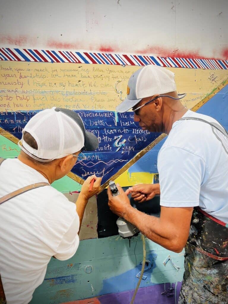 Two artists work side by side on a large, colorful painting filled with handwritten messages and bold geometric shapes.