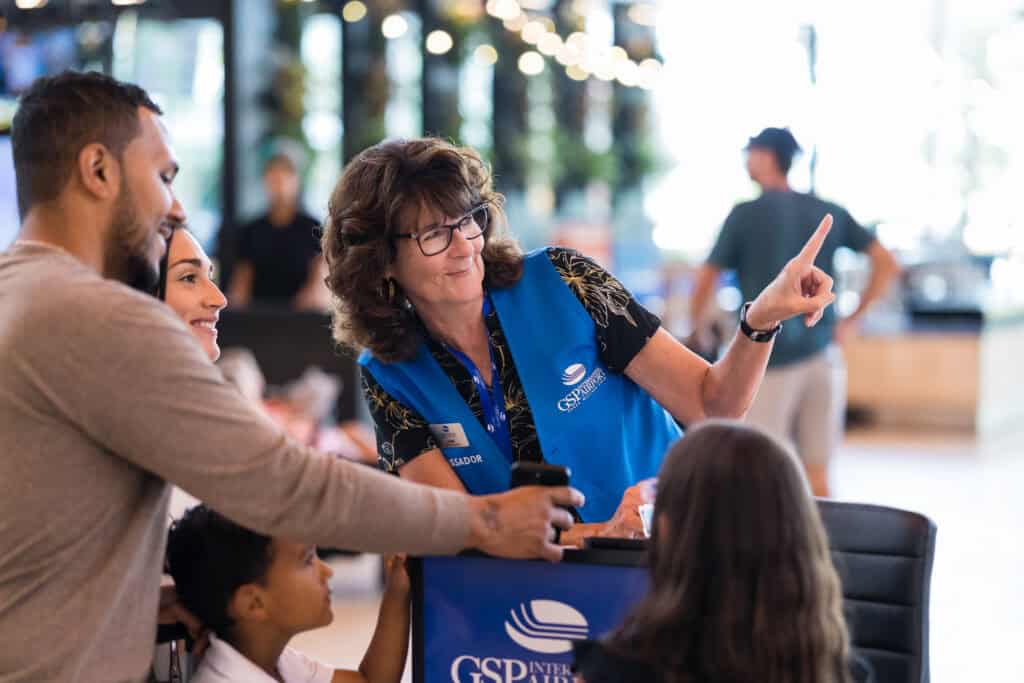 A smiling GSP Airport ambassador wearing a blue vest assists a family at an information cart, pointing in a direction while the family, including two children, listens attentively.