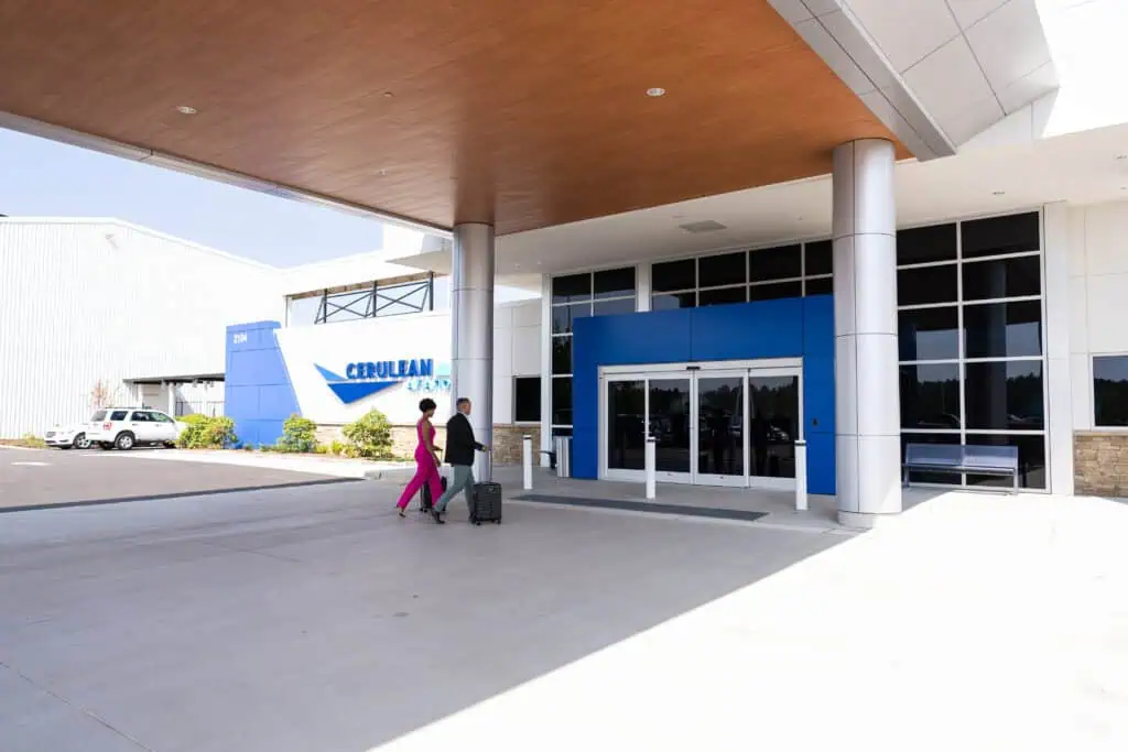wo people walk toward the entrance of the Cerulean Aviation terminal, pulling luggage, under a covered drop-off area. The modern building features large glass windows and a blue accent around the doors. A sign displaying "Cerulean Aviation" and the address "2104" is visible on the left. wo people walk toward the entrance of the Cerulean Aviation terminal, pulling luggage, under a covered drop-off area. The modern building features large glass windows and a blue accent around the doors. A sign displaying "Cerulean Aviation" and the address "2104" is visible on the left