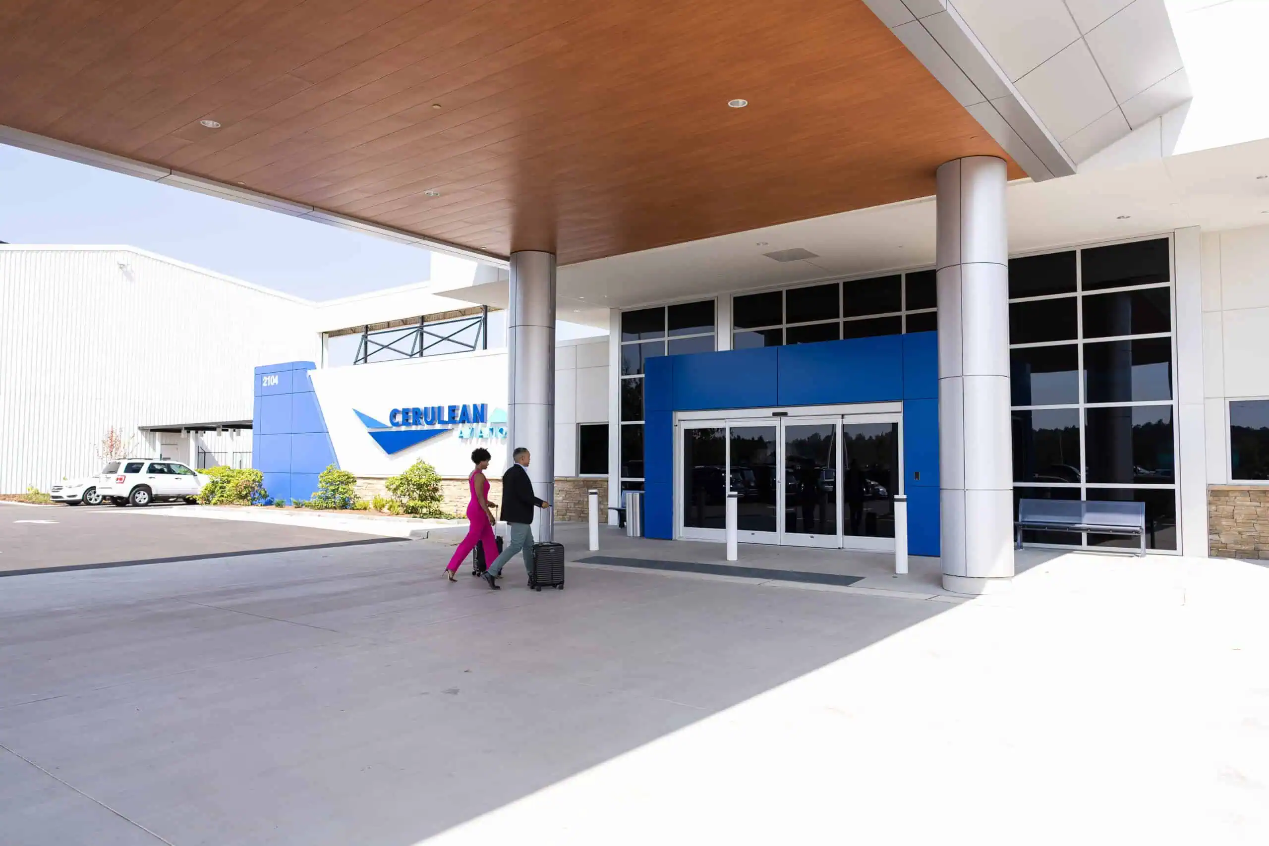 wo people walk toward the entrance of the Cerulean Aviation terminal, pulling luggage, under a covered drop-off area. The modern building features large glass windows and a blue accent around the doors. A sign displaying "Cerulean Aviation" and the address "2104" is visible on the left. wo people walk toward the entrance of the Cerulean Aviation terminal, pulling luggage, under a covered drop-off area. The modern building features large glass windows and a blue accent around the doors. A sign displaying "Cerulean Aviation" and the address "2104" is visible on the left