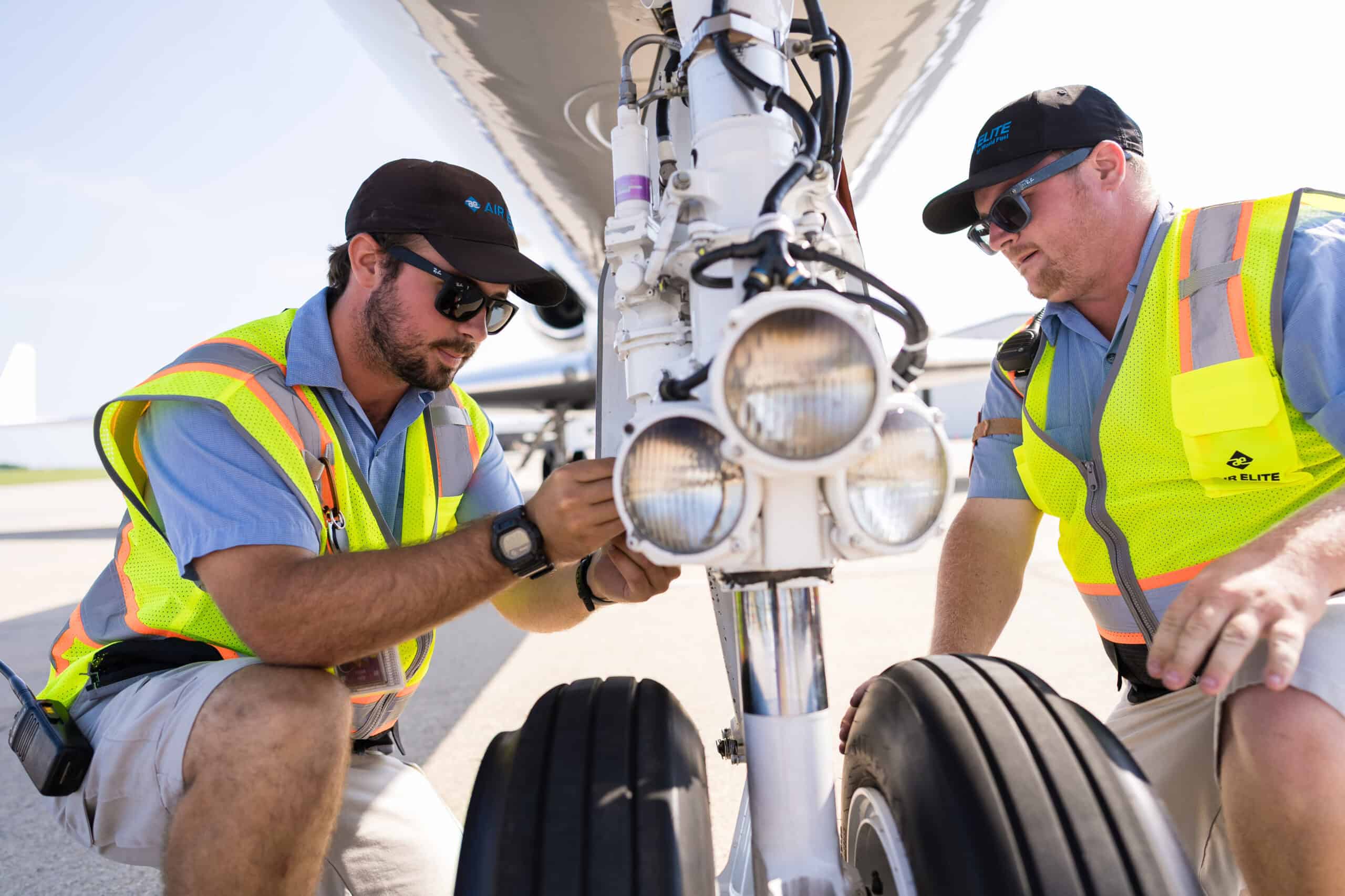 Two ground crew members wearing safety vests and caps inspect the nose landing gear of an aircraft on the tarmac, with focus on the aircraft lights and wheels.